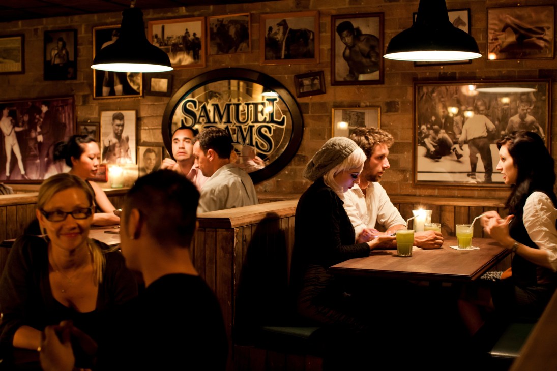 The Baxter Inn interior with patrons at wooden tables, framed photographs on walls, and Samuel Smith's beer signage.