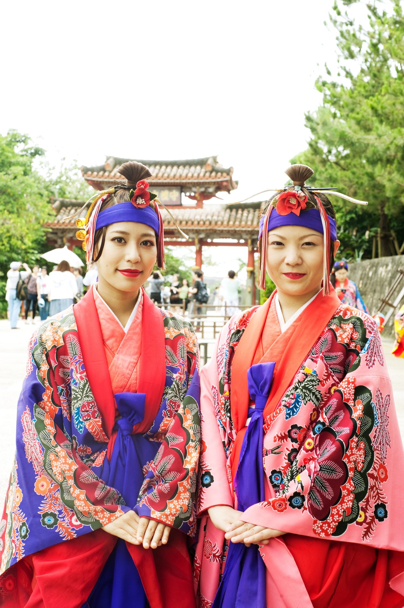 Two women wearing traditional Okinawan Ryuso ethnic costumes with colorful embroidered jackets, red skirts, and blue headbands at Shurijo castle.