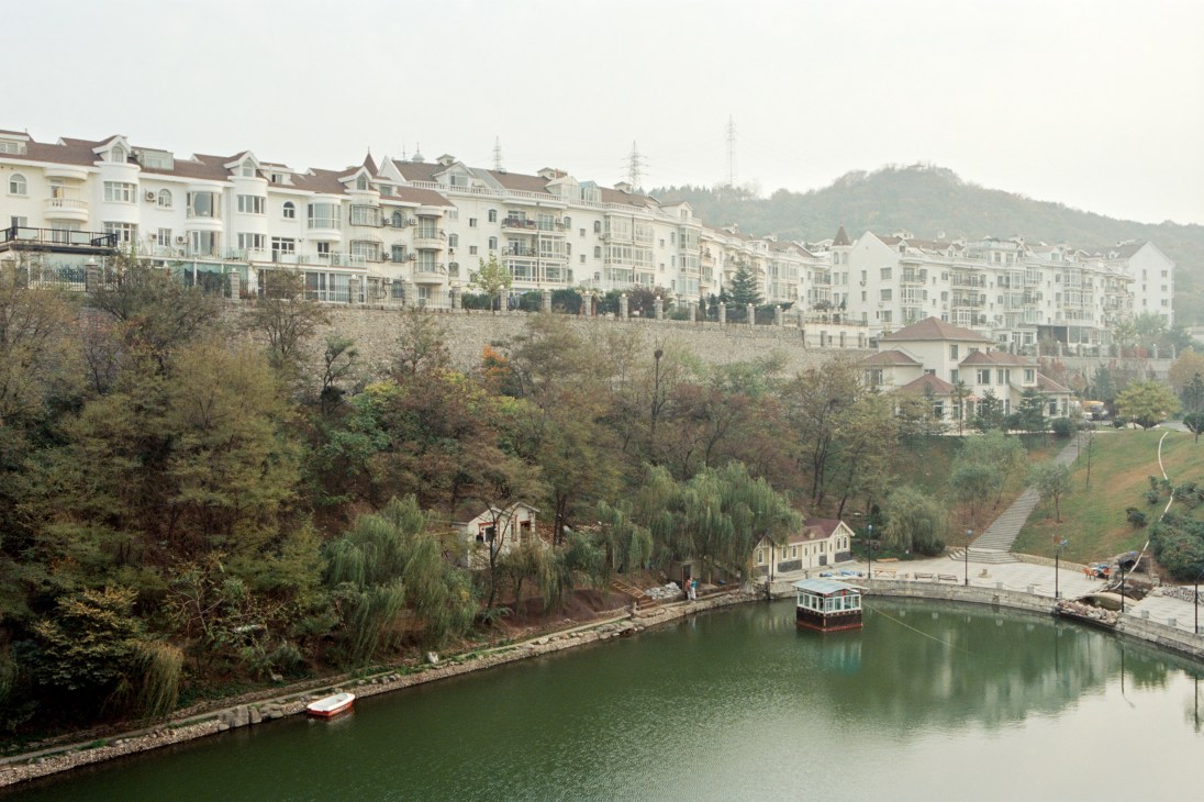 Gated residential community with cream-colored apartment buildings overlooking a river and wooded hillside in Zhongshan, Dalian.