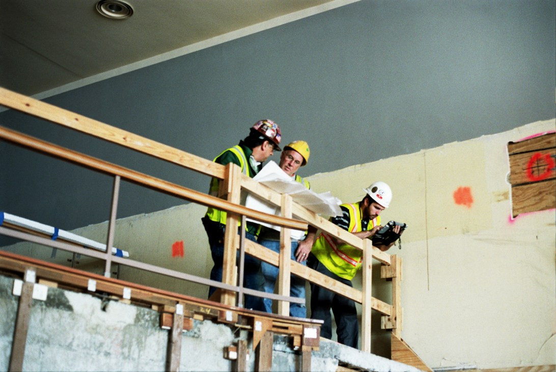 Three workers in hard hats and high-visibility vests stand on wooden scaffolding during the Trusteeship Council Chamber restoration.