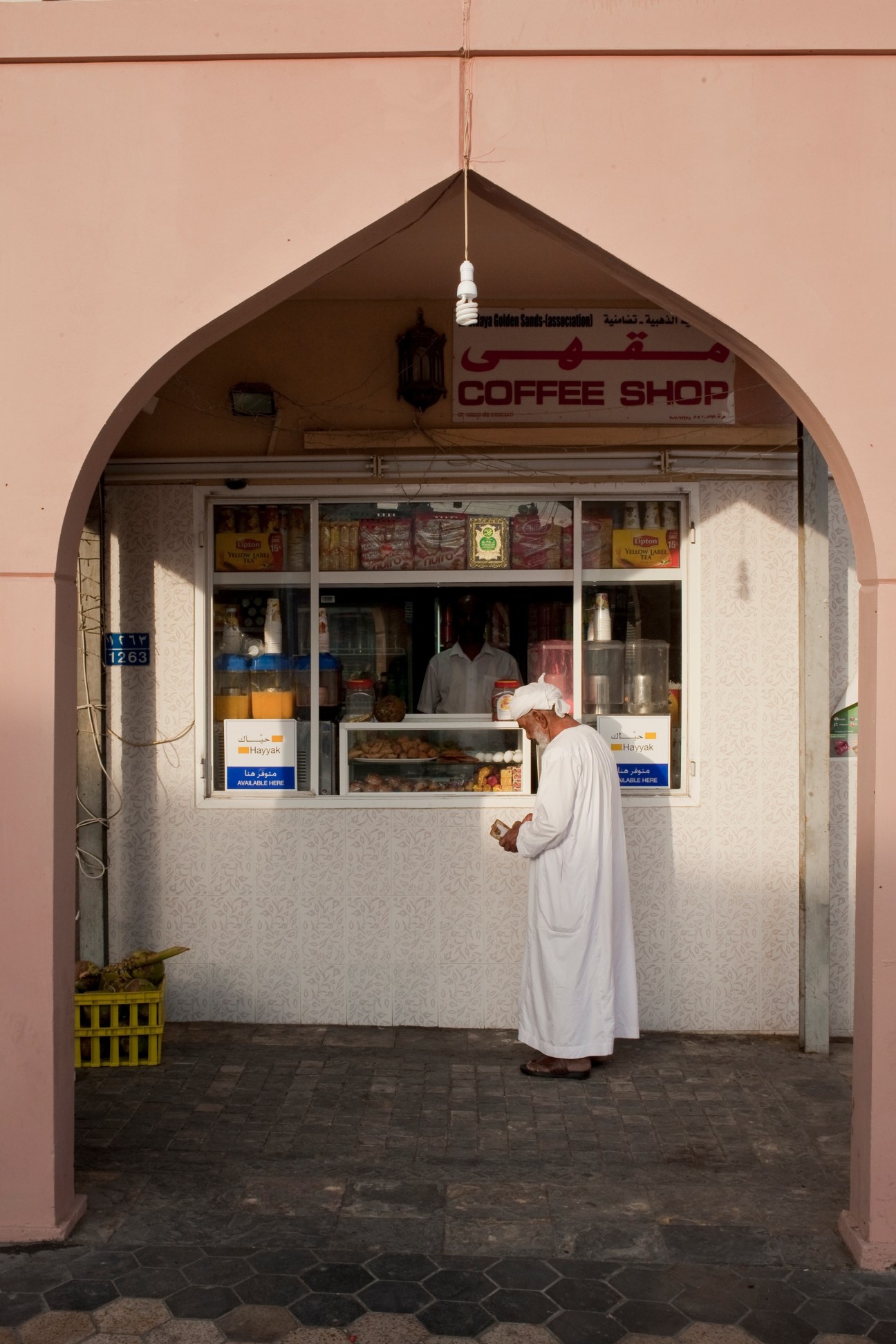 Man in white traditional dress standing at a convenience shop counter in Muscat with pink arched doorway.