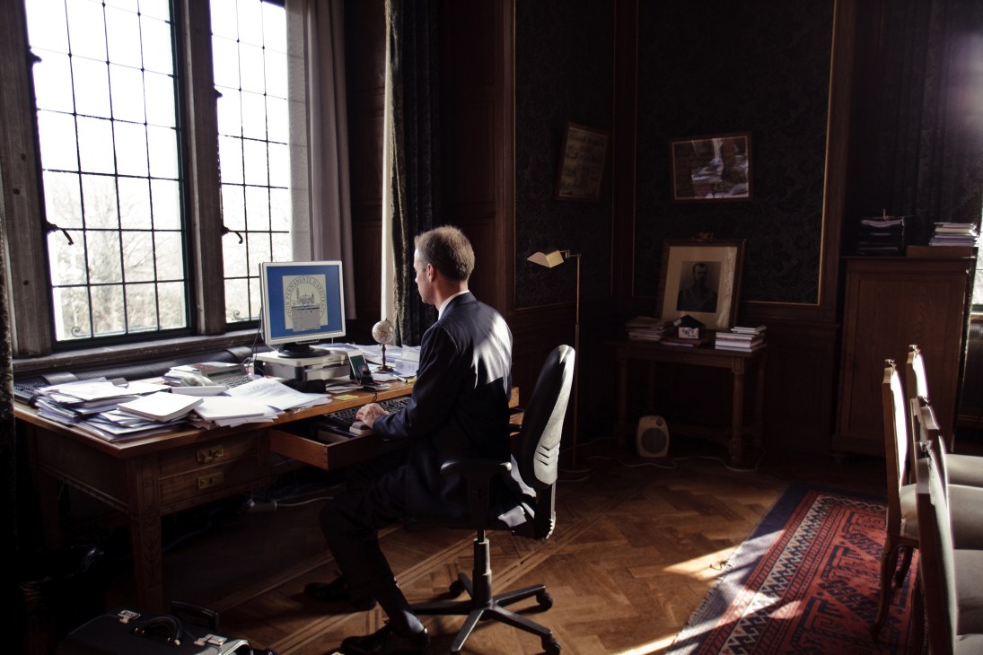 Man in business suit sitting at wooden desk in ornate office with papers and computer monitor.