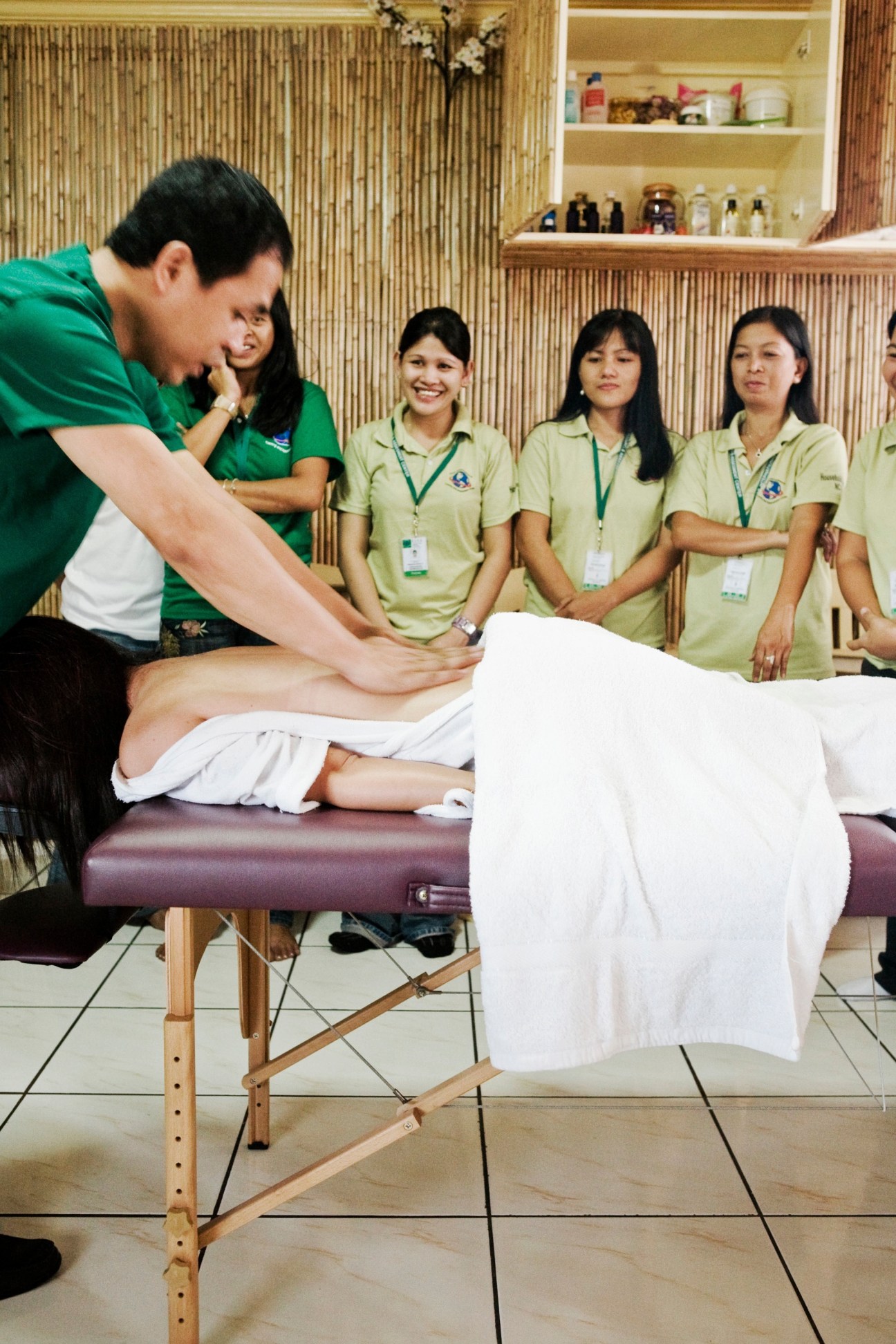 Massage instructor demonstrates technique on student while trainees in green uniforms observe in classroom.