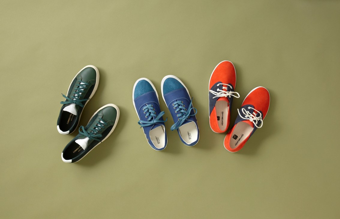 Three pairs of spring trainers displayed on a sage green background: Common Projects from New York in dark green with white sole, Gram Shoes from Stockholm in blue with white trim, and Veja from Paris in orange with white laces.