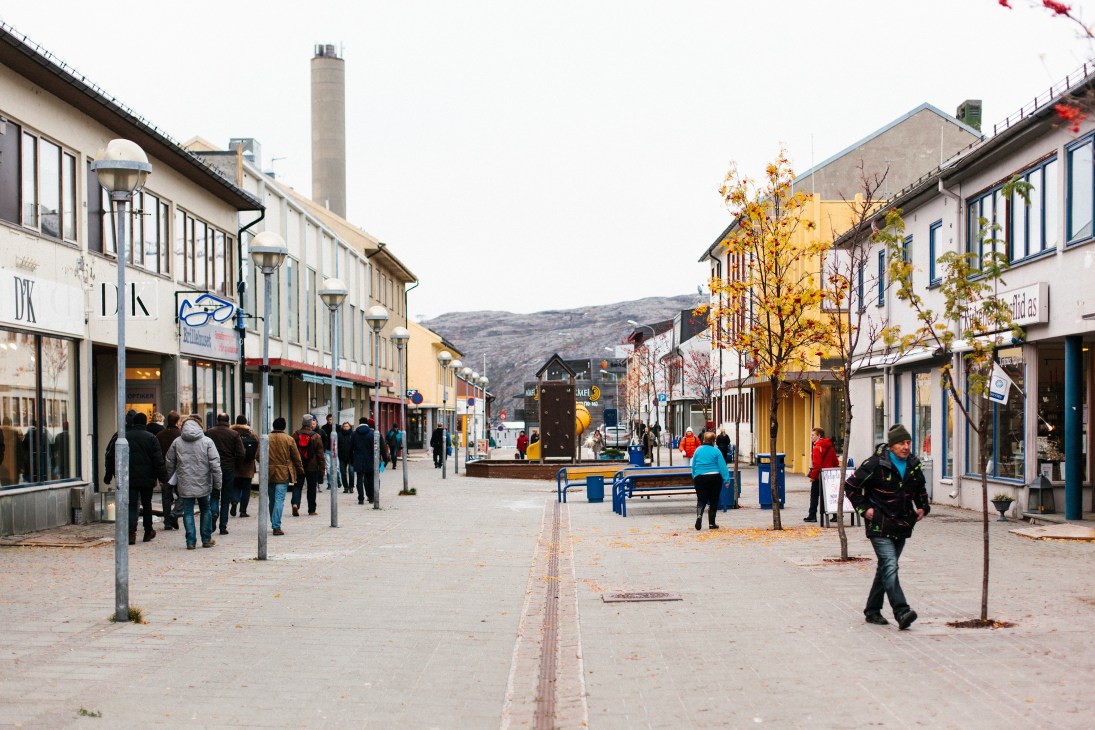 Central Kirkenes pedestrian zone with colorful storefronts, shoppers, and mountains in background.