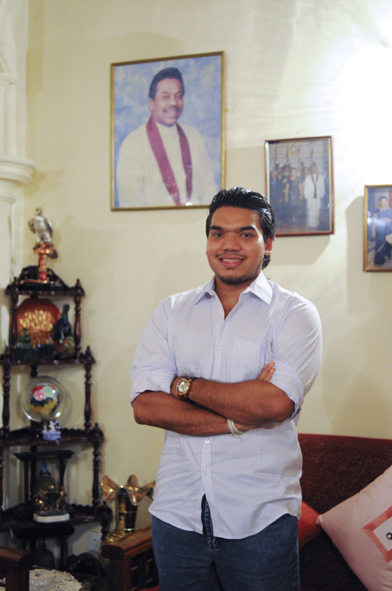 Namal Rajapaksa, a young man in a light blue shirt, stands with arms crossed smiling at the camera in an office with framed portraits on the wall behind him.