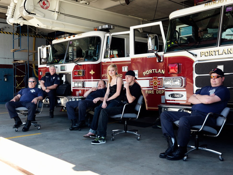Portland fire department members and a woman posing in front of red and white fire trucks in a station garage.