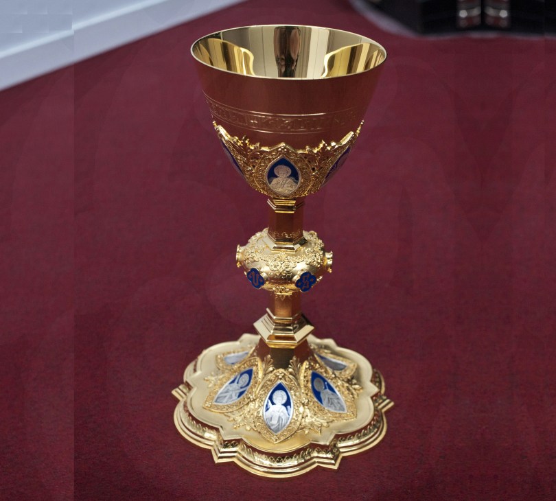 Ornate golden chalice with blue enamel details and religious medallions on burgundy background.