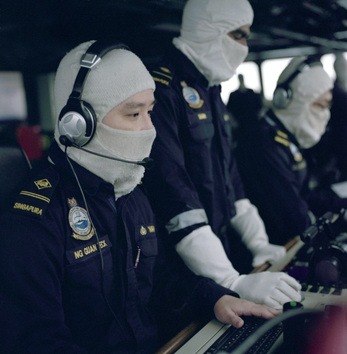 Singapore Navy personnel in uniform and headsets monitor equipment on a ship's bridge.