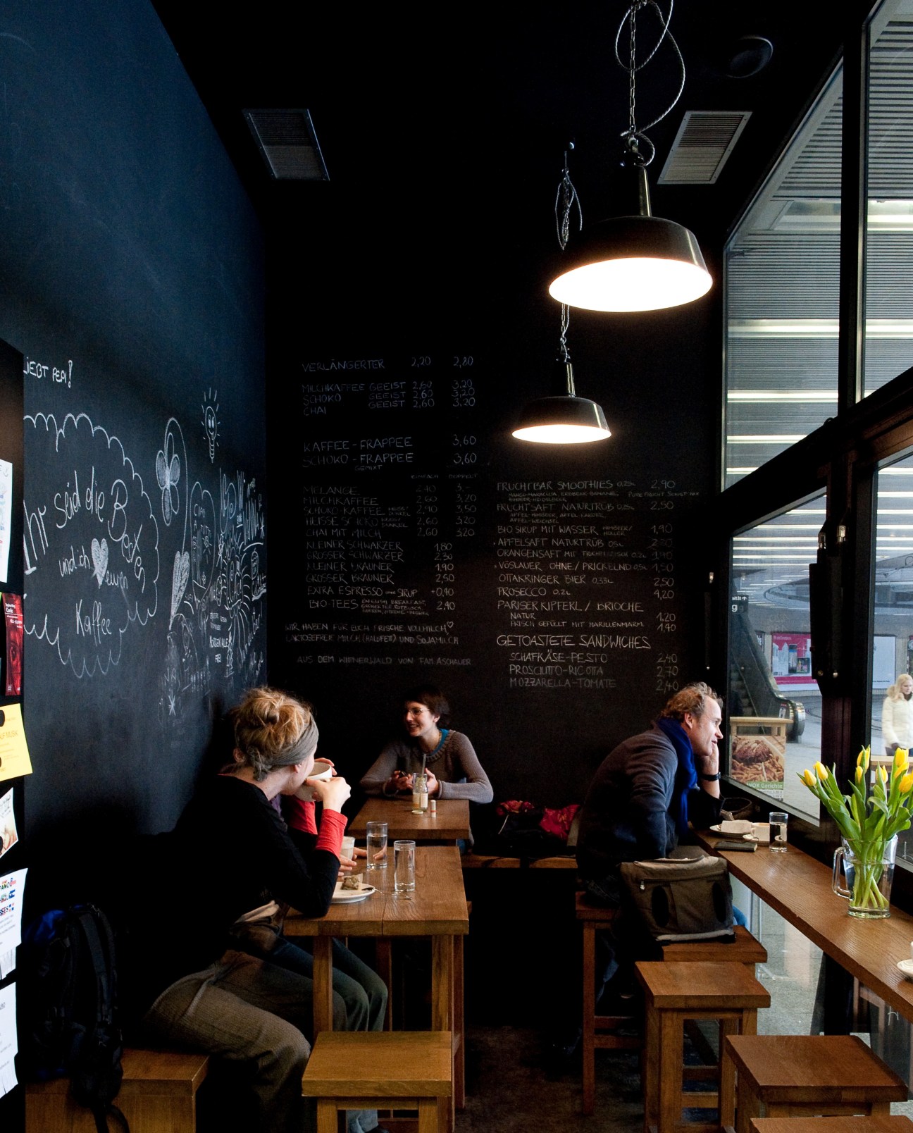 Customers seated at wooden tables inside Kaffeeküche café with chalkboard menu and hanging pendant lights.