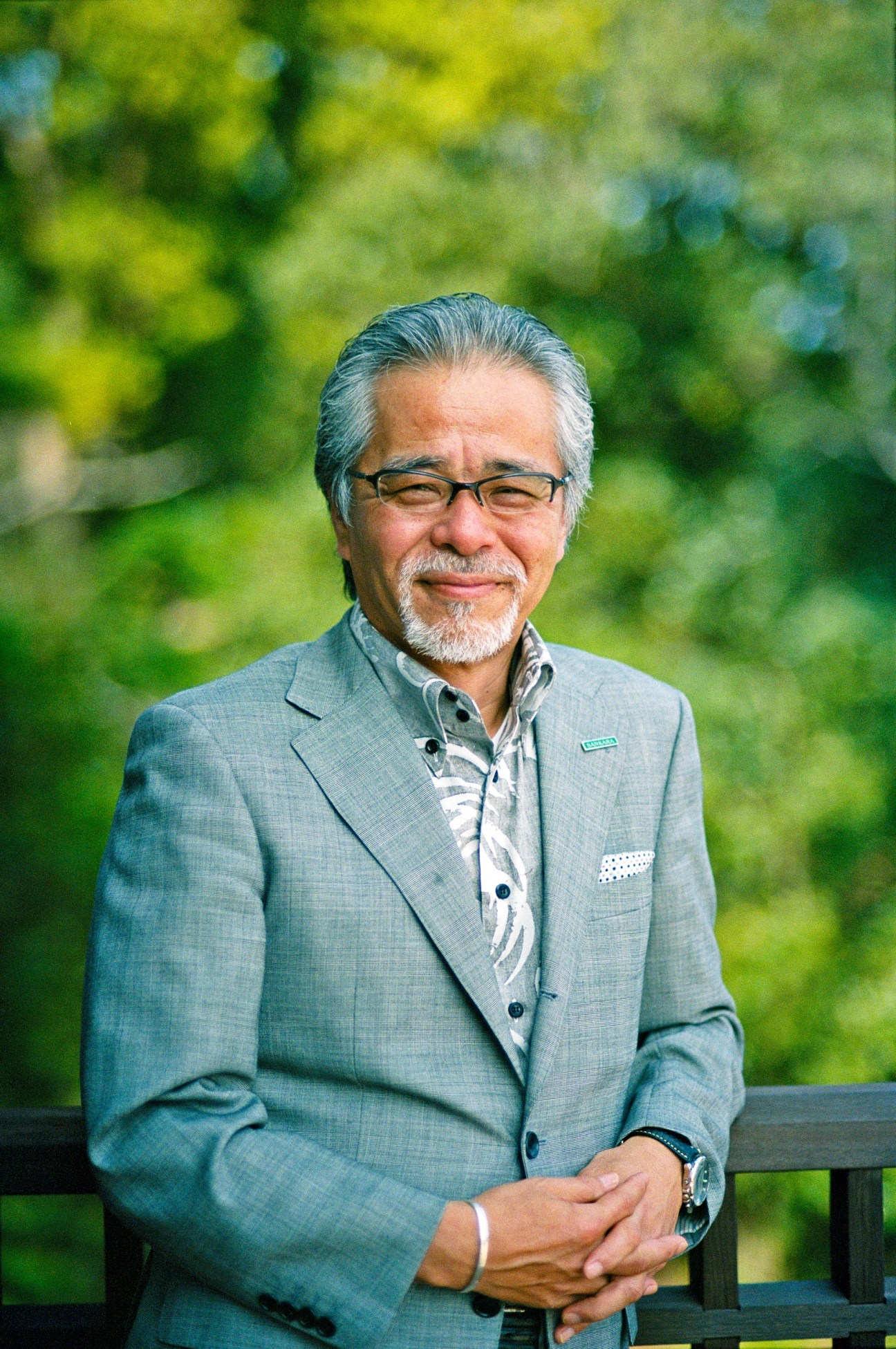 Jiro Sato, designer of Sankara Hotel & Spa, poses outdoors in a grey blazer against a blurred forest background.