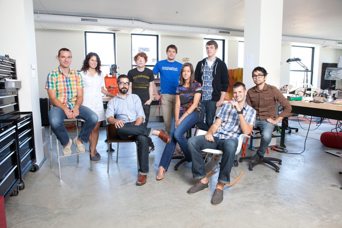 Team of ten people posing together in a modern workshop space with industrial tooling and equipment visible.