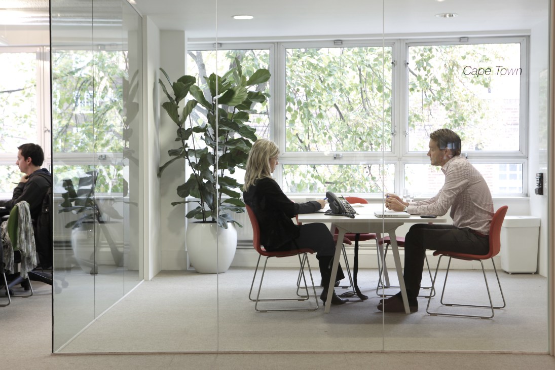 Two people sit at a table in a modern glass-walled meeting room with views of trees outside and the text 'Cape Town' visible on the glass.