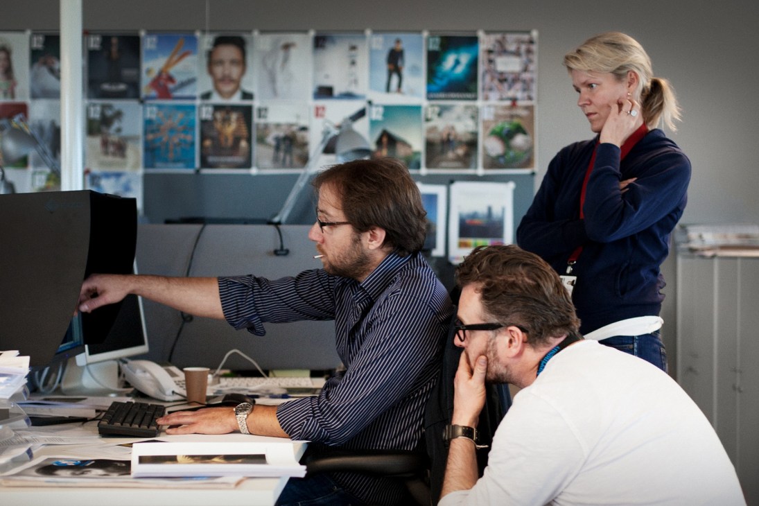 Staff members discussing a story in a newspaper office with photo layouts displayed on wall behind them.