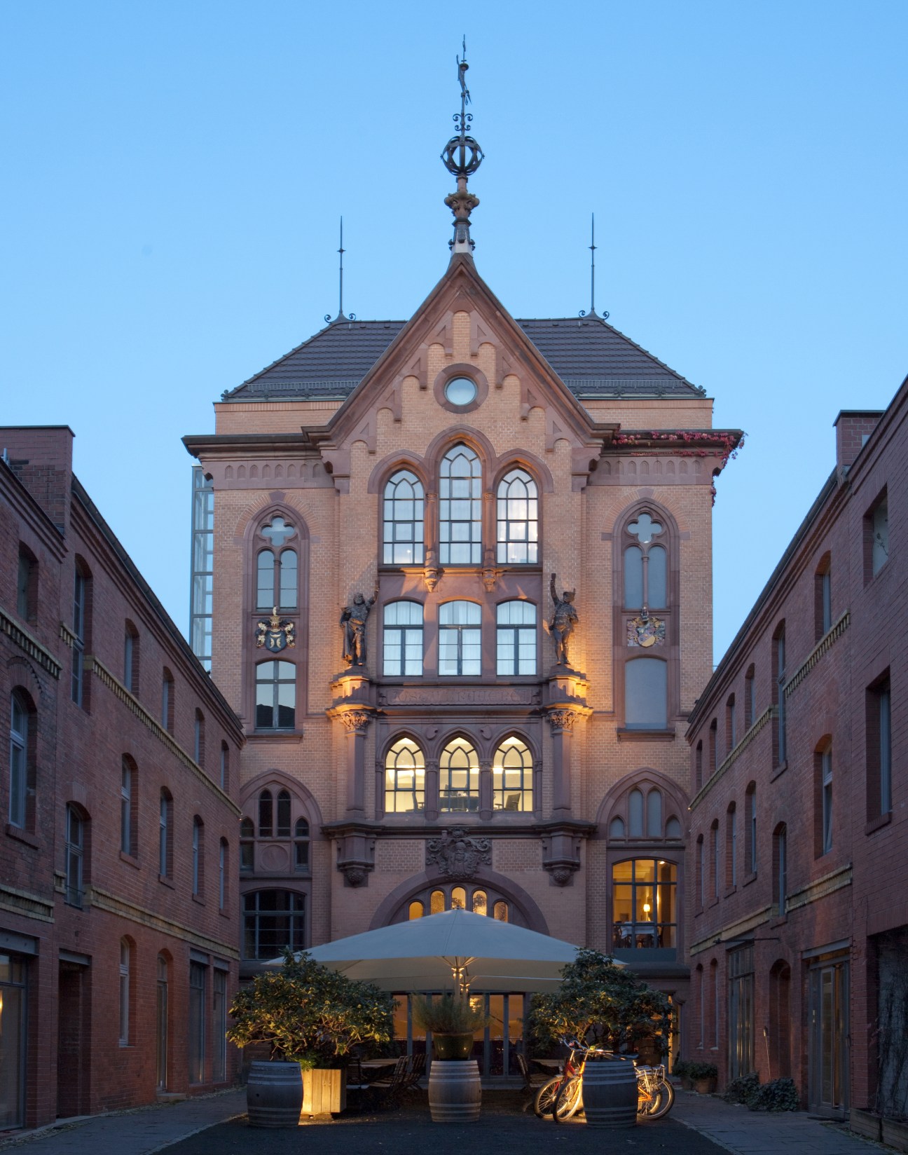Red brick Neo-Gothic building courtyard with illuminated windows at dusk, featuring a white umbrella and outdoor seating area.