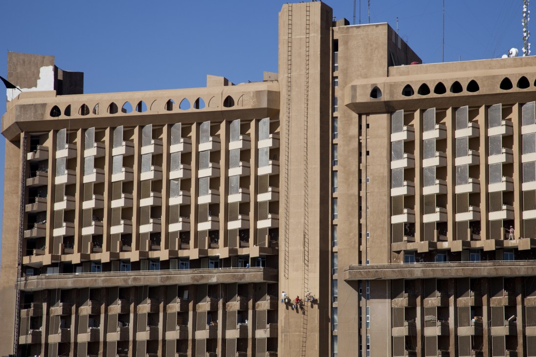 Workers on scaffolding at the Mansoor Media Hotel, a large beige brutalist building with distinctive architectural features.