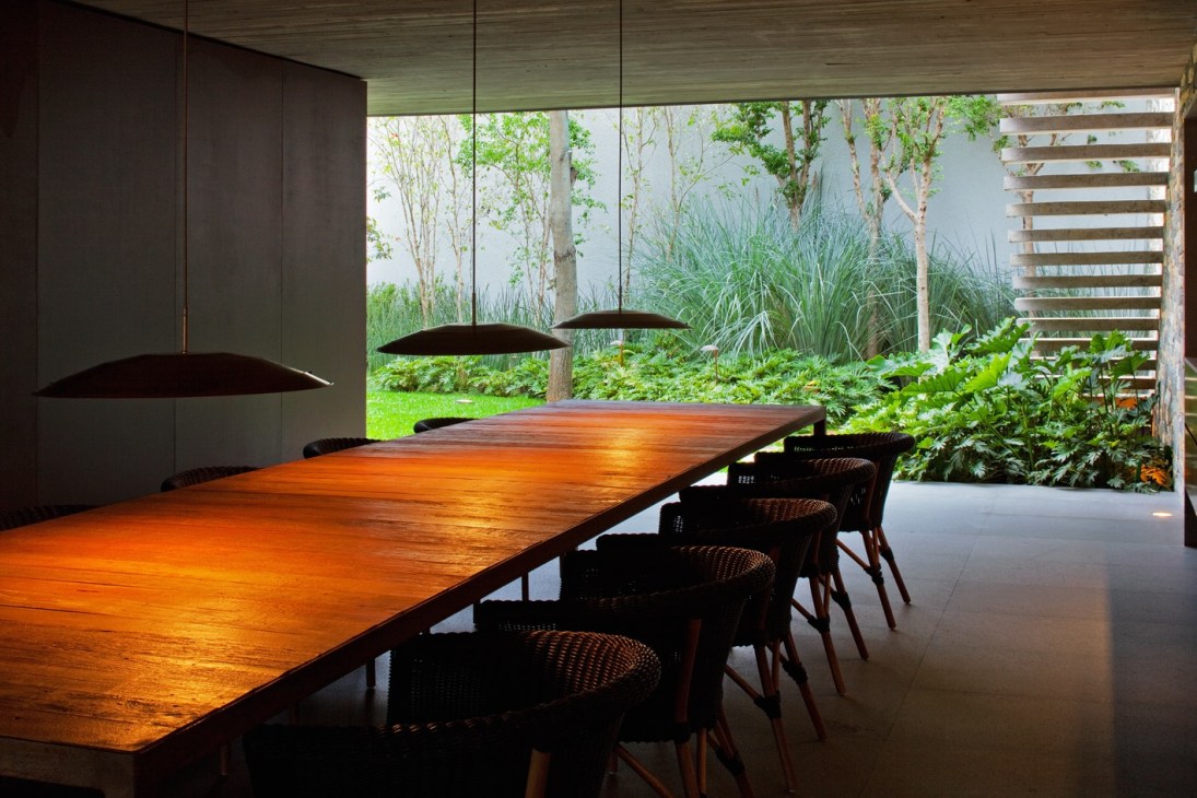Marcio Kogan-designed wooden dining table with black chairs and pendant lights overlooking a garden.