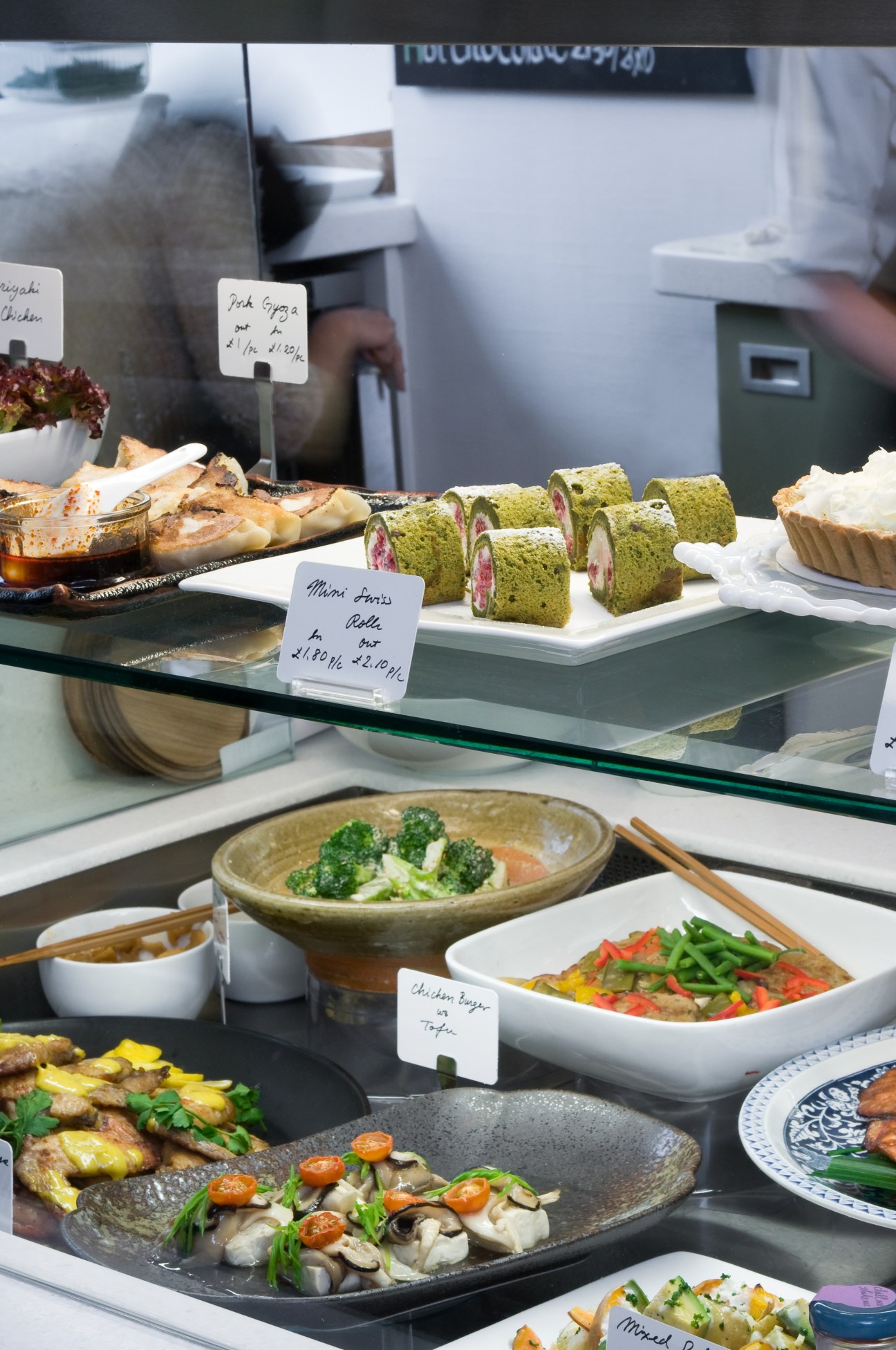Japanese deli display at Tombo restaurant showing plated dishes with price labels and matcha cakes.
