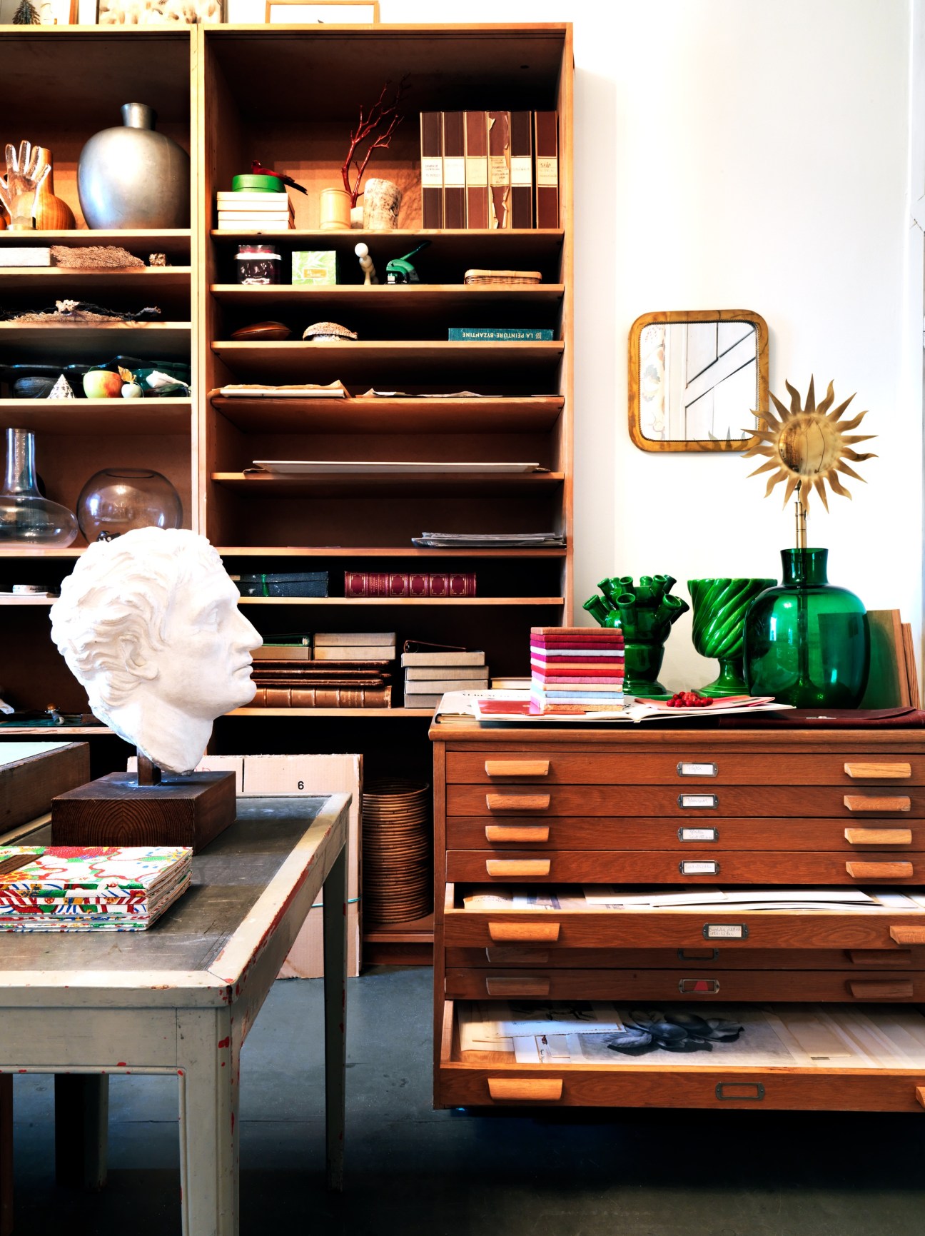 Recreated office display featuring wooden shelving, desk with drawers, white bust sculpture, green glass vessels, and curated decorative objects.