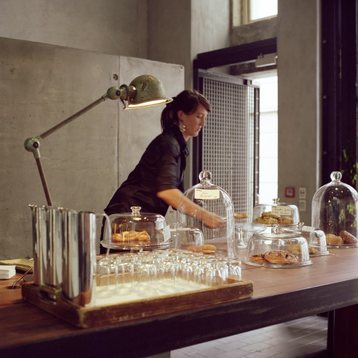 Woman arranging pastries under glass domes at a counter in a modern café with industrial lighting.