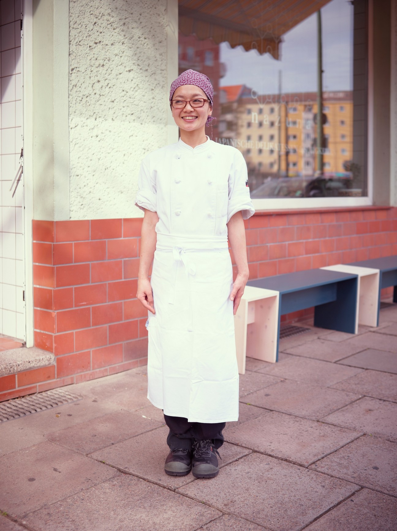 Smiling chef in white chef's coat and pink headwrap standing outside Nazuna delicatessen in Berlin.
