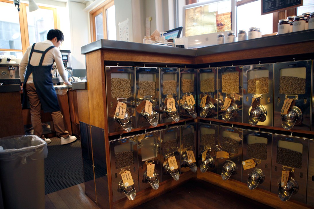 Coffee shop interior with barista at counter and display cases of coffee beans in labeled dispensers.