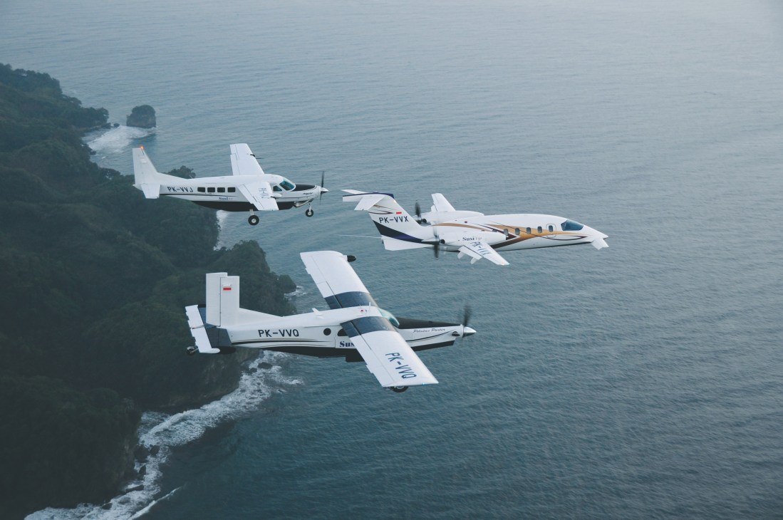 Three white Susi Air propeller planes flying in formation over Indonesian coastline with dense jungle and blue ocean.