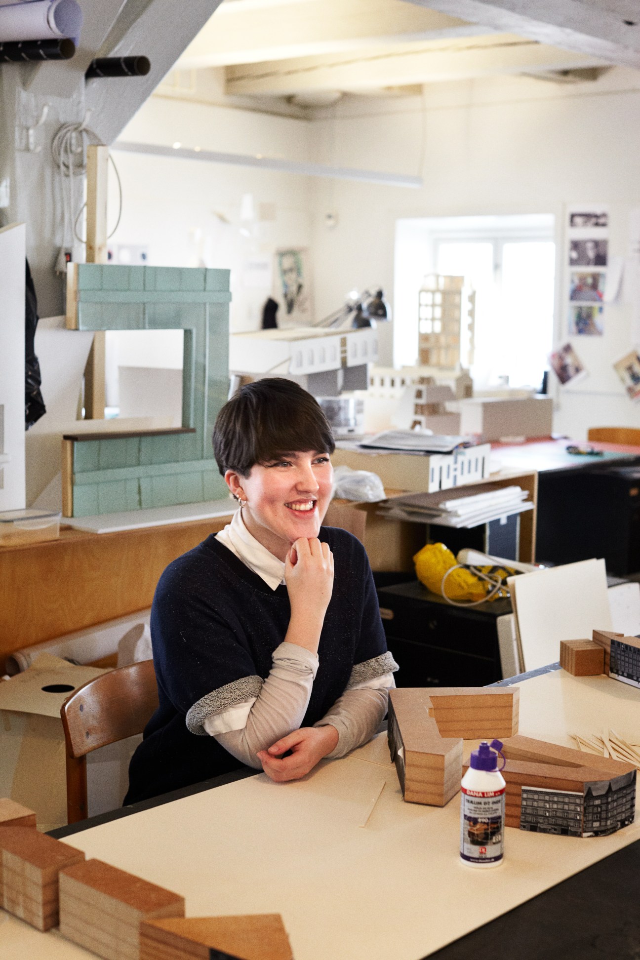 Ebba Tham smiles at her desk in an architecture studio surrounded by scale models and design materials.
