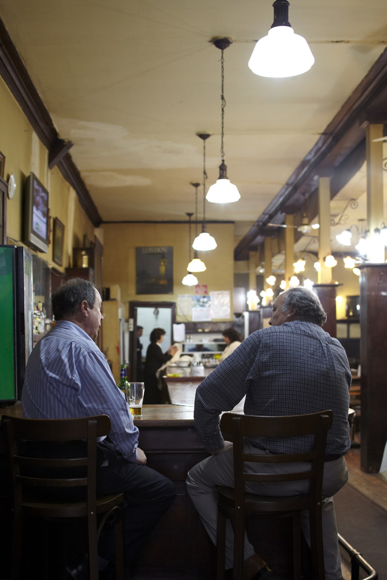 Two men sit at the bar counter of Bar Inglés in Valparaíso, with pendant lights hanging above.