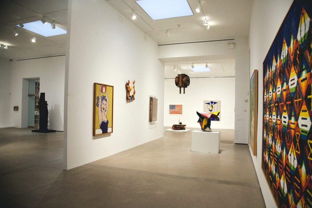 Contemporary art gallery interior displaying paintings, sculptures and mixed media works on white walls under skylights.