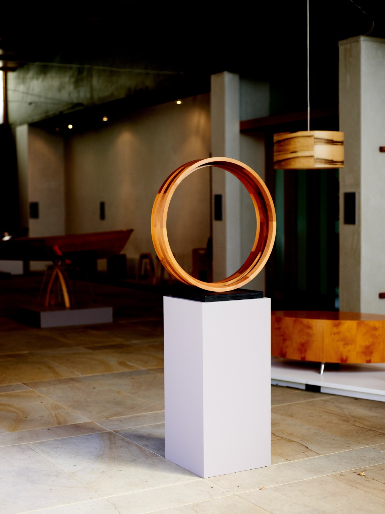 A polished wooden circular form mounted on a white pedestal in a gallery space with other wooden artworks visible in the background.