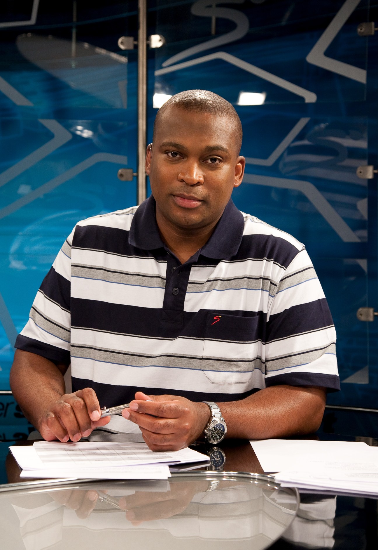 Robert Marawa, a broadcaster in a striped polo shirt, sits at a desk in a television studio.