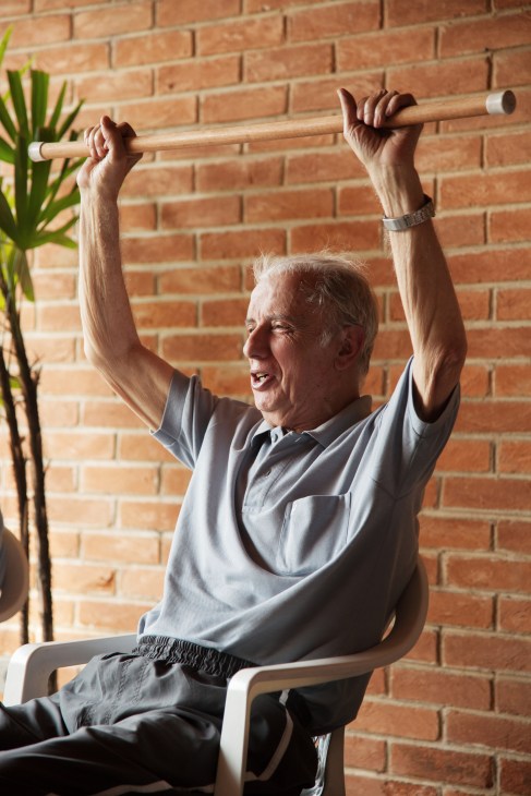 An elderly man performs an overhead exercise with a wooden bar while seated against a brick wall.