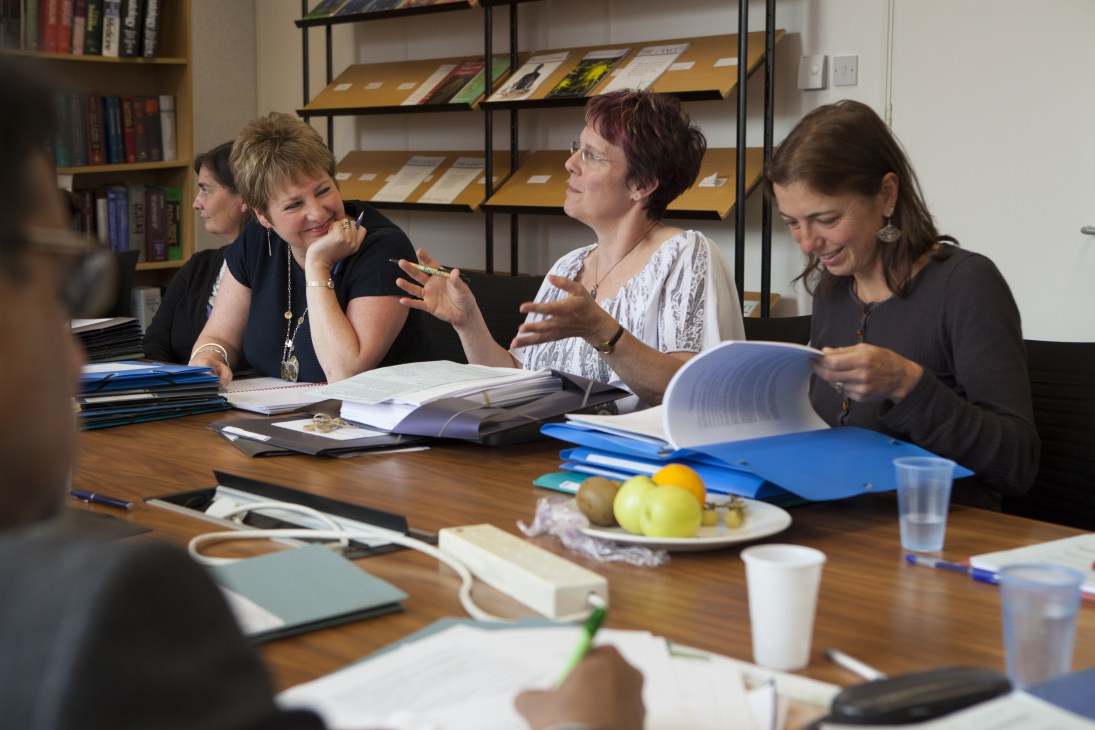 Four women seated at a wooden table during The Lancet's weekly peer-review meeting, reviewing documents and journals.