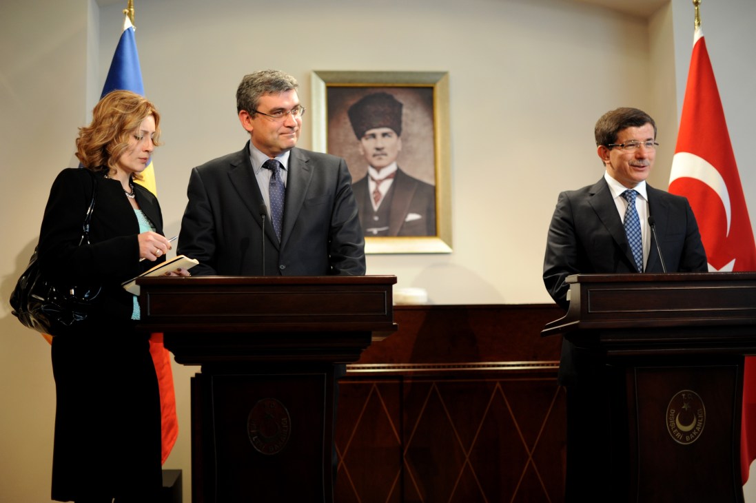 Davutoglu stands at a podium during a press conference with the Romanian minister of foreign affairs, with flags and a portrait of Ataturk in the background.