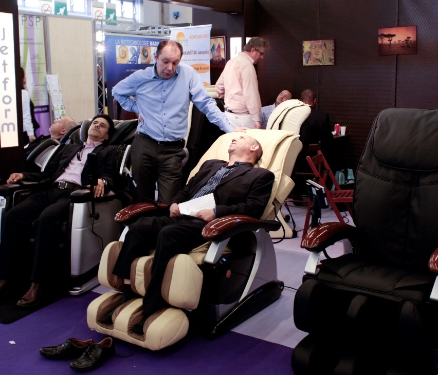 Trade fair visitors testing reclining massage chairs at Pharmagora pharmaceutical exhibition.