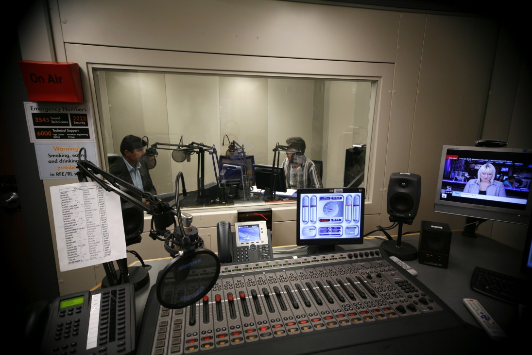 Recording studio with two broadcasters at microphones behind glass, control desk with mixer visible in foreground.