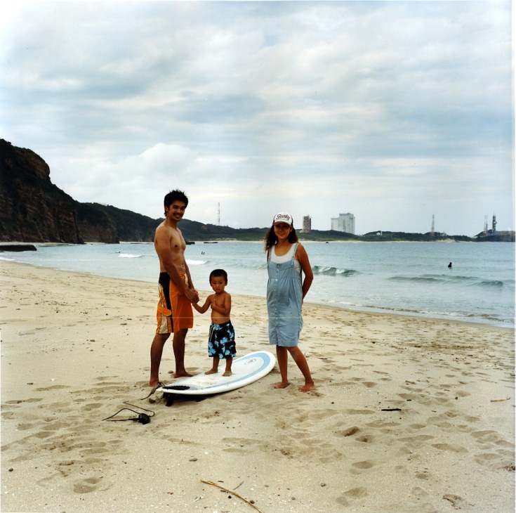 Family of four with surfboard on a beach with space center facilities visible in the background.