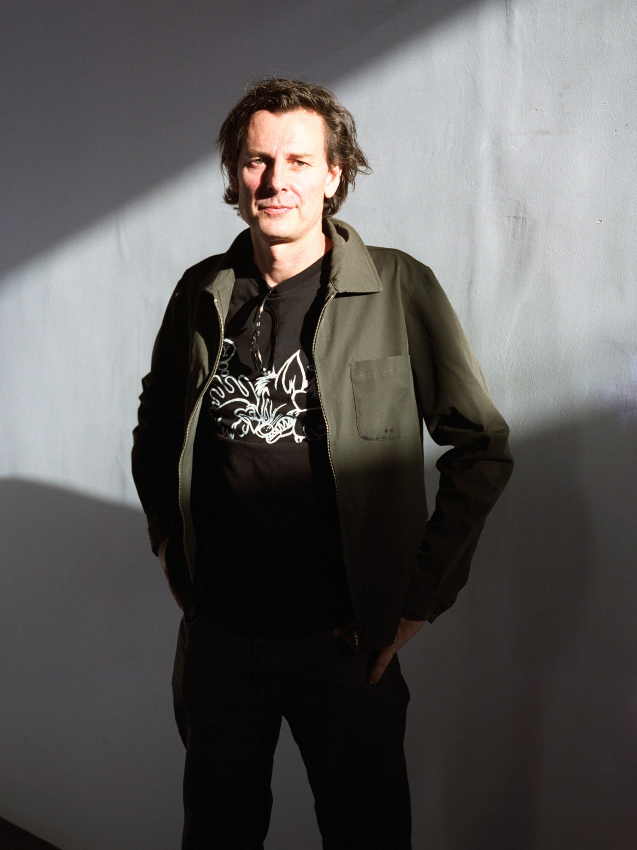 Portrait of James Brearley, an Australian architect, standing against a white wall wearing a dark jacket and patterned shirt.