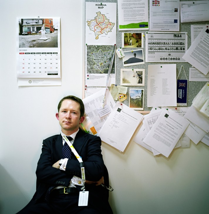 Man in dark suit with credential lanyard stands against wall covered with Kosovo maps, documents and photographs.