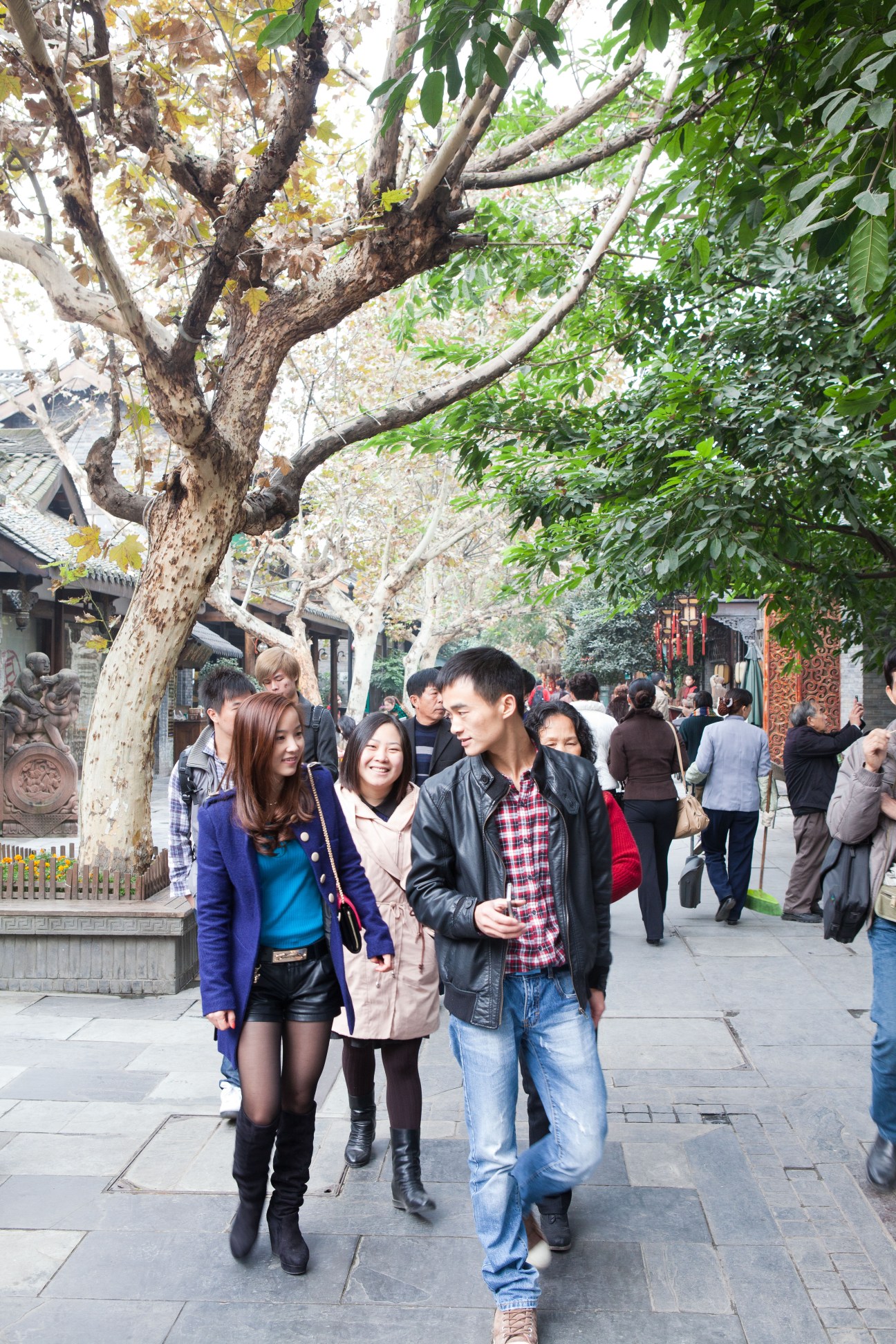 Three young people walk together under a large tree in a pedestrian shopping area with historic buildings.