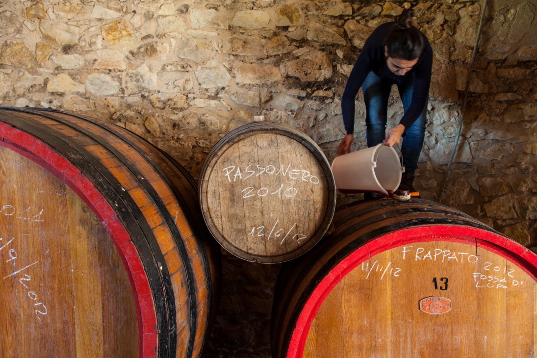 Woman pouring wine from a container into wooden barrels labeled with vintage dates in a stone cellar.