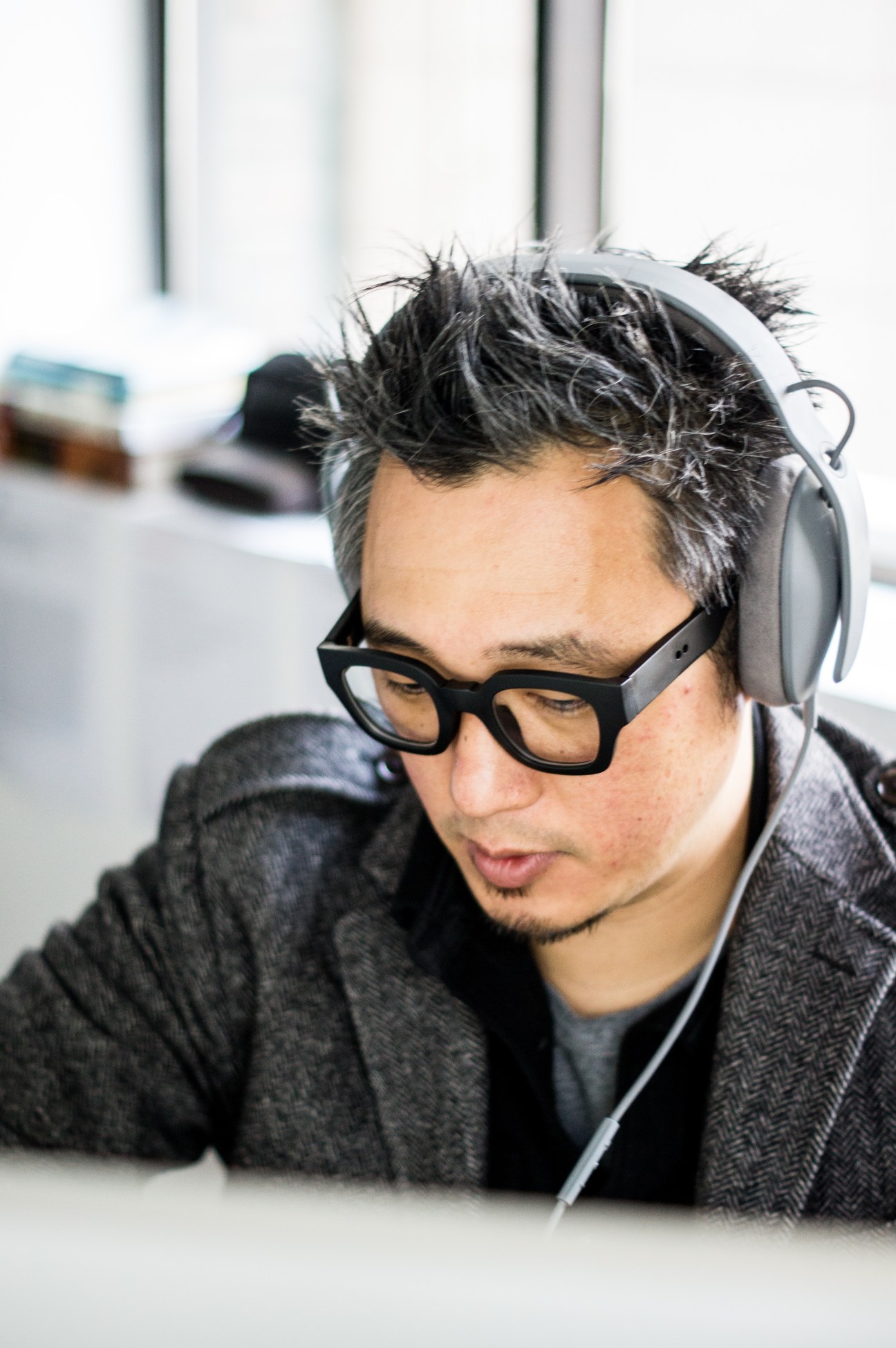 Bernie Cho wearing black-framed glasses and white headphones around his neck, looking downward in an office setting.