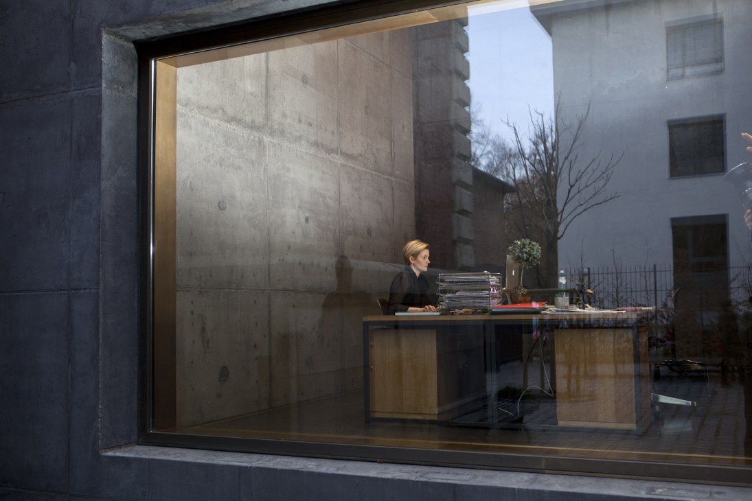Woman at desk inside modern office space viewed through large window with concrete interior walls.