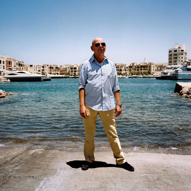 Walter Plangenauer stands on a beach in Aqaba wearing sunglasses, a light blue shirt and khaki pants, with beachfront buildings and boats visible behind him.