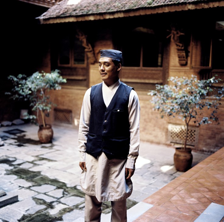 A waiter in traditional Nepali dress stands in a temple house hotel courtyard.