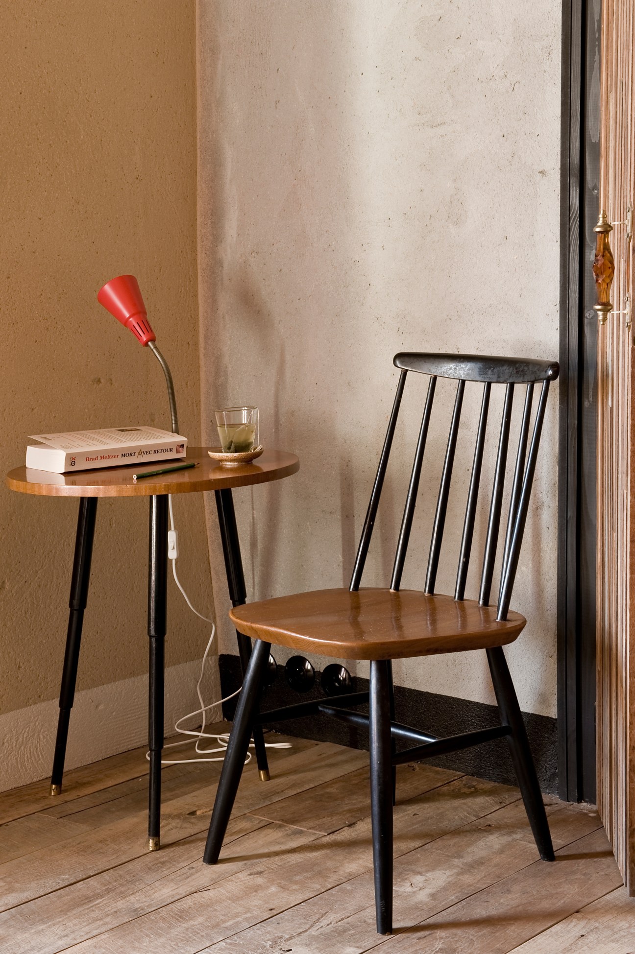 A mid-century modern wooden chair with black spindle back sits next to a round side table holding books, a red desk lamp, and a glass, against a textured plaster wall.