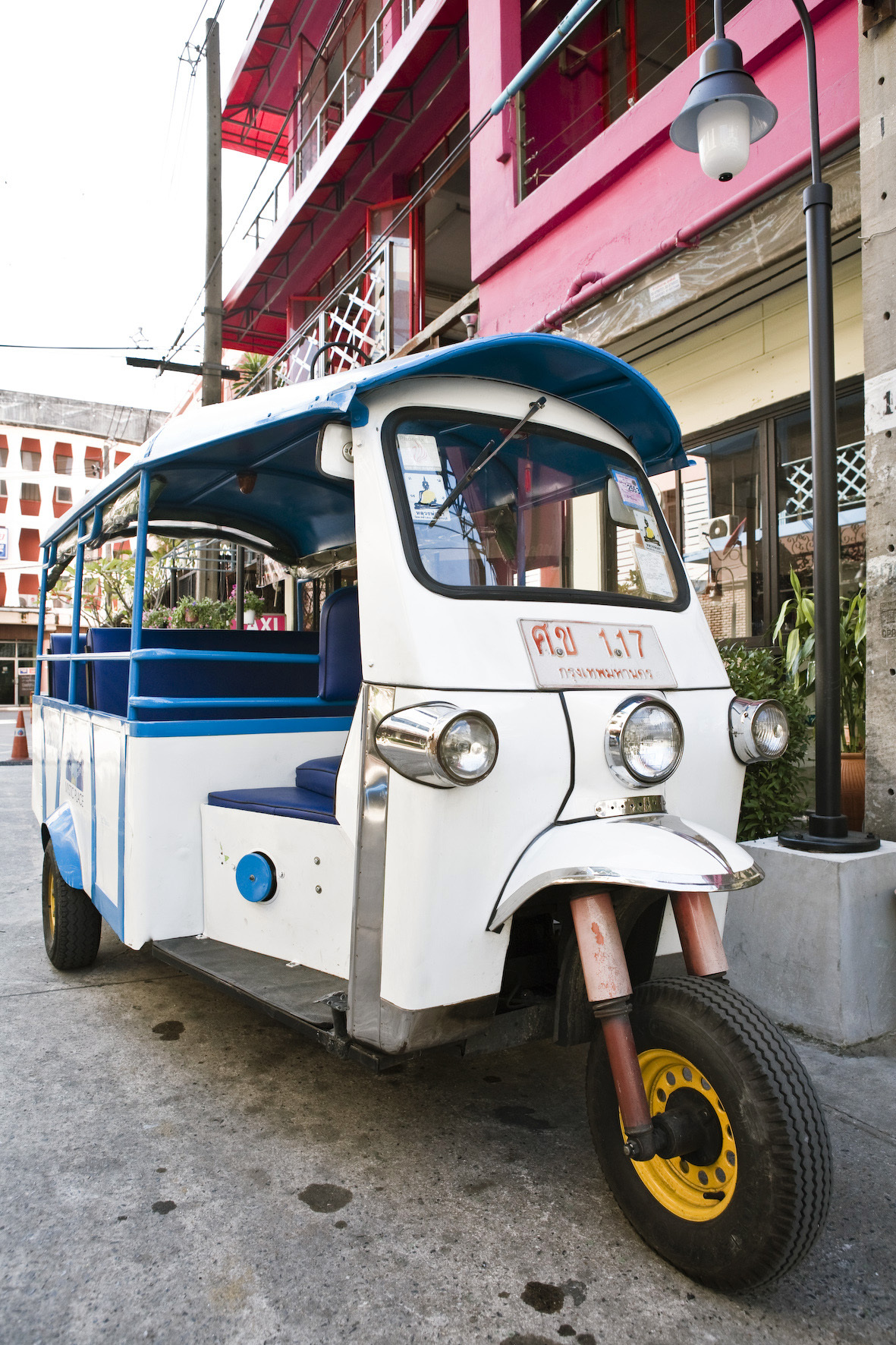 White and blue tuktuk parked on a Bangkok street with colorful buildings in background.