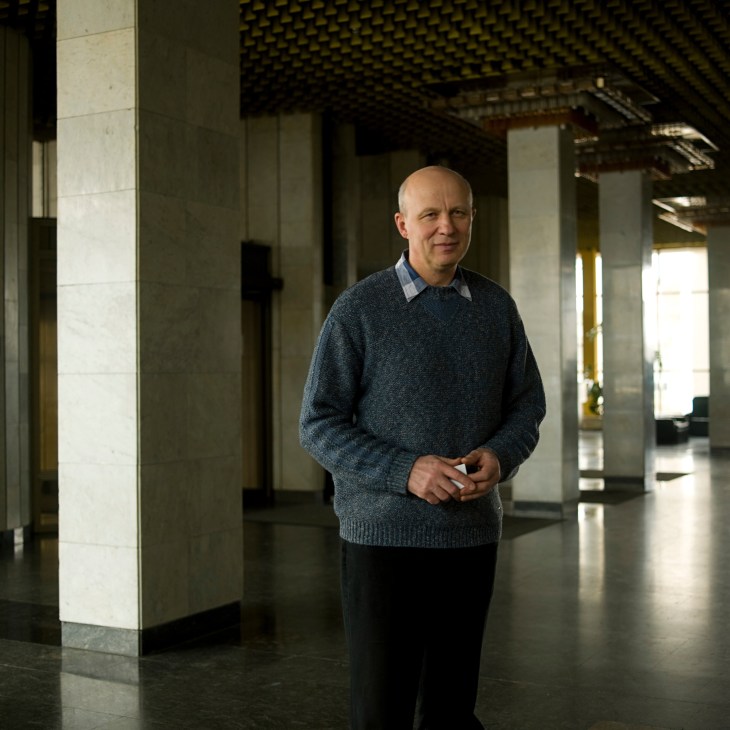 Bald man in grey sweater standing in modern interior with white columns.