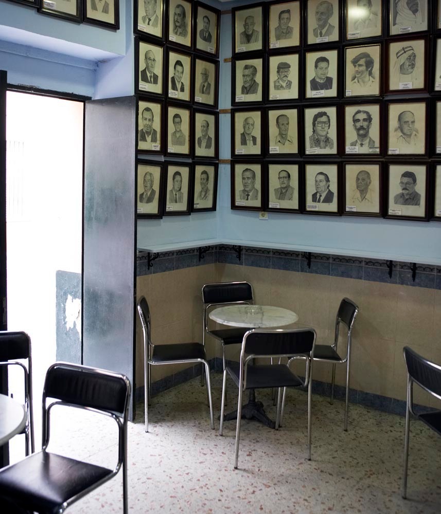 Bar interior with framed portrait drawings of singing competition winners covering the walls above a small round table and chairs.